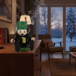 Räuchermännchen Mini „Bergmann“ (8 cm), farbig gestaltet mit grüner Bergmannsmütze, Werkzeug und schwarzer Tracht, rauchend auf einem Tisch in gemütlicher Wohnatmosphäre vor winterlicher Fensterkulisse dekoriert.