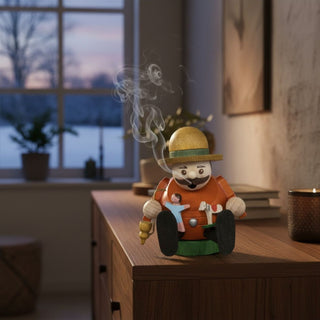 Räuchermännchen Mini „Spielzeughändler“ (8 cm), farbig gestaltet mit Hut und kleinen Holzfiguren in den Händen, rauchend auf einer Kommode in gemütlicher Wohnatmosphäre vor winterlicher Fensterkulisse dekoriert.