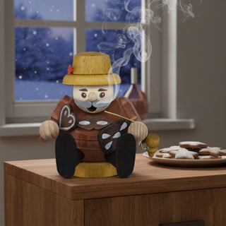 Räuchermännchen Mini „Lebkuchenhändler“ (8 cm), farbig gestaltet mit Hut und Lebkuchenherzen in der Hand, rauchend auf einer Kommode neben Plätzchen vor winterlicher Fensterkulisse dekoriert.