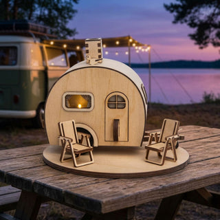 äucherwohnwagen „KWALM“ (ca. 15 cm) aus naturbelassenem Holz, detailreich gestaltet mit Tür, Fenster und kleinem Schornstein, aus dem Rauch aufsteigt. Steht auf einem Holztisch mit zwei Mini-Liegestühlen, im Hintergrund ein Campingplatz am See bei Abenddämmerung mit Wohnwagen und Lichterkette.