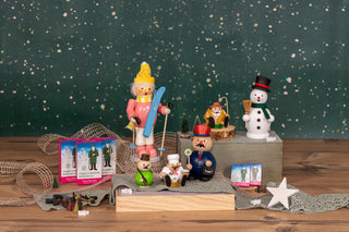 Das Bild zeigt eine festliche Weihnachtsszene mit verschiedenen weihnachtlichen Figuren und Dekorationen. Es gibt drei Räuchermännchen, die jeweils unterschiedliche Gegenstände halten: einer hält Skier, ein anderer eine Trompete und der dritte einen kleinen Baum. Außerdem gibt es eine Schneemannfigur mit schwarzem Hut und rotem Schal, die auf der rechten Seite des Bildes steht. Der Hintergrund zeigt eine grüne Kulisse mit weißen Sprenkeln, die Schneeflocken ähneln. Vor der Szene sind drei  Schachteln Räuche