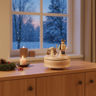 Spieluhr „Förster“ (ca. 12 cm) aus naturbelassenem Holz, mit drehbarer Szene eines Försters neben einem kleinen Handwagen und Tannenbäumen. Die Spieluhr spielt die Melodie „Oh Tannenbaum“. Sie steht auf einem Holzmöbel in einer gemütlichen Wohnstube; im Hintergrund ein großes Fenster mit verschneiter Winterlandschaft sowie Kerzenlicht und Tannenzweig, die eine warme, festliche Atmosphäre schaffen.