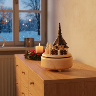 Spieluhr „Seiffener Kirche“ (ca. 21 cm) aus naturbelassenem Holz, mit detailreicher Darstellung der Seiffener Kirche, umgeben von kleinen Figuren und Tannenbäumen. Die drehbare Szene wird von der Melodie „Oh Tannenbaum“ begleitet. Die Spieluhr steht auf einem Holzmöbel in einer gemütlichen Wohnstube; im Hintergrund ein Fenster mit verschneiter Winterlandschaft sowie eine Kerze und ein Adventskranz, die eine warme, festliche Atmosphäre schaffen.