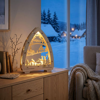LED-Lichterspitze „Wildfütterung“ aus naturfarbenem Holz (29 cm) mit warmweißer LED-Beleuchtung. Das mehrlagige, filigran ausgeschnittene Motiv zeigt eine winterliche Waldszene mit Rehen an einer Futterraufe, umgeben von Tannenbäumen und einer verschneiten Landschaft. Im Hintergrund sind ein Dorf mit Kirche und Fachwerkhäusern sowie fliegende Vögel dargestellt. Die dekorative Lichterspitze steht auf einer Holzkommode in einem gemütlichen Wohnraum vor einem Fenster mit Blick auf ein verschneites Winterdorf.