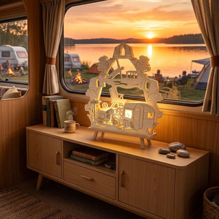 LED-Standleuchte „(N)Ostalgie Camping“ aus naturfarbenem Holz (40 cm) mit warmweißer LED-Beleuchtung. Das detailreich ausgeschnittene Motiv zeigt einen Wohnwagen, ein Moped, Campingmöbel, Bäume und einen Pavillon in einer idyllischen Campinglandschaft. Die dekorative Leuchte steht auf einem Sideboard im Inneren eines Wohnwagens. Durch das Fenster ist ein Campingplatz am See mit Zelten, Wohnwagen, Lagerfeuern und einem stimmungsvollen Sonnenuntergang zu sehen. 