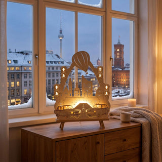 LED-Standleuchte „(N)Ostalgie Trabant und Wartburg“ aus naturfarbenem Holz (44 cm) mit warmweißer LED-Beleuchtung. Das detailreich ausgeschnittene Motiv zeigt einen Trabant und einen Wartburg auf einer Straße vor einer Stadtkulisse mit Fernsehturm, historischen Gebäuden und Heißluftballon. Ampelsymbole im Stil der bekannten Ost-Ampelmännchen rahmen die Szene dekorativ ein. Die Leuchte steht auf einer Kommode vor einem Fenster mit Blick auf eine winterliche Stadtansicht.