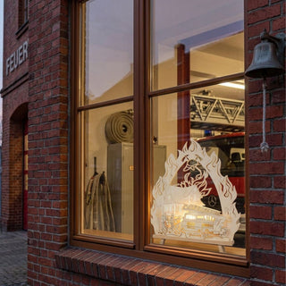 LED-Standleuchte „Feuerwehr“ aus naturfarbenem Holz (43 cm) mit warmweißer LED-Beleuchtung. Das detailreich ausgeschnittene Motiv zeigt ein Feuerwehrauto mit Drehleiter im Einsatz vor einem brennenden Haus, umrahmt von stilisierten Flammen. Die dekorative Leuchte steht in einem Fenster einer Feuerwache, im Hintergrund sind Feuerwehrfahrzeug, Ausrüstung und Backsteinfassade sichtbar. Die warme Beleuchtung setzt das Feuerwehrmotiv eindrucksvoll in Szene und sorgt für eine stimmungsvolle Atmosphäre.