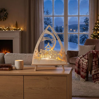 LED-Standleuchte „Weihnachtsmann“ aus naturfarbenem Holz (39 cm) mit warmweißer LED-Beleuchtung. Die dekorative Leuchte in spitz zulaufender Form zeigt den Weihnachtsmann mit Geschenkesack und Schlitten, begleitet von einem Rentier. Über der winterlichen Szene sind ein geschmückter Weihnachtsbaum, Sterne und ein Komet mit Schweif filigran ausgeschnitten. Die Leuchte steht auf einem Holztisch in einem festlich dekorierten Wohnraum vor einem Fenster mit Blick auf eine verschneite Winterlandschaft. 