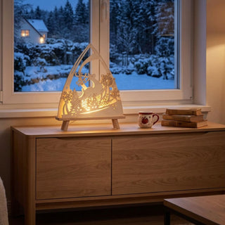 LED-Standleuchte „Winterzauber“ aus naturfarbenem Holz (39 cm) mit warmweißer LED-Beleuchtung. Die dekorative Leuchte in spitz zulaufender Form zeigt ein springendes Rentier in einer winterlichen Landschaft mit filigran gestalteten Schneeflocken, Bäumen und glitzernden Sternelementen. Die Leuchte steht auf einem Sideboard vor einem Fenster mit Blick auf einen verschneiten Garten und einen winterlichen Nadelwald. 
