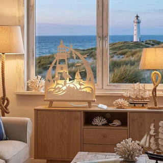 LED-Standleuchte „Küstenliebe“ aus naturfarbenem Holz (41 cm) mit warmweißer LED-Beleuchtung. Das filigran ausgeschnittene Motiv zeigt einen Leuchtturm, Möwen, Wellen und ein Segelboot in einer maritimen Küstenszene. Die dekorative Leuchte steht auf einem Holzsideboard vor einem Fenster mit Blick auf Dünen, Meer und einen Leuchtturm. Maritime Dekorationselemente wie Korallen und Segelschiffe unterstreichen die gemütliche Küstenatmosphäre.