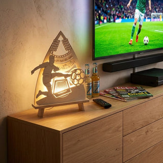 LED-Standleuchte „Fußballfreunde“ aus naturfarbenem Holz (41 cm) mit warmweißer LED-Beleuchtung. Das filigran ausgeschnittene Motiv zeigt einen Fußballspieler beim Schuss auf ein Tor, umgeben von Stadiondetails und Flutlichtmasten. Die dekorative Leuchte steht auf einem Holzsideboard neben einem Fernseher mit Fußballübertragung und sorgt für eine stimmungsvolle Atmosphäre für Fußballfans.