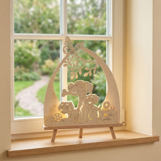 LED-Standleuchte „Hundefreund“ aus naturfarbenem Holz (41 cm) mit warmweißer LED-Beleuchtung. Das filigran gearbeitete Motiv zeigt einen ausgewachsenen Hund mit zwei Welpen unter einem Ast mit Schmetterling. Blumen, Gras und ein Spielball ergänzen die liebevolle Szene. Die dekorative Leuchte steht auf einer Fensterbank vor einem Fenster mit Blick ins Grüne und sorgt für eine gemütliche, wohnliche Atmosphäre.