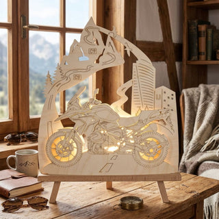 LED-Standleuchte „Motorradfreund“ aus naturfarbenem Holz (41 cm) mit warmweißer LED-Beleuchtung. Das detailreich ausgeschnittene Motiv zeigt ein Reiseenduro-Motorrad auf einer kurvenreichen Straße. Berggipfel, Serpentinen, Nadelbäume und eine moderne Stadtkulisse mit Hochhaus rahmen die Szene ein. Die dekorative Leuchte steht auf einem Holztisch vor einem Fenster mit Bergblick und schafft mit ihrem warmen Licht eine gemütliche Atmosphäre für Motorrad- und Reiseliebhaber.