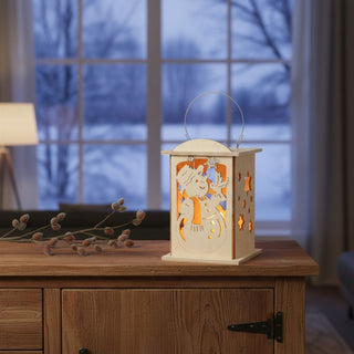 Laterne für 1 Teelicht oder mit LED‑Stripe „Schneemann“, 16 cm, aus naturbelassenem Holz mit filigraner Schneemann‑Darstellung und Sternen, dekorativ auf einem Holzsideboard mit Zweigen platziert, im Hintergrund ein gemütliches Wohnzimmer mit Sessel und großer Fensterfront mit winterlicher Landschaft, stimmungsvoll beleuchtet durch warmes Licht im Inneren der Laterne.