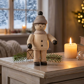 Miniatur „Junge mit Laterne“ (ca. 5 cm) aus naturbelassenem Holz, mit kleiner Figur in Winterkleidung und einer Laterne in der Hand. Die Figur steht auf einem Holzmöbel in einer gemütlichen Stube; im Hintergrund ein Fenster mit verschneiter Winterlandschaft, eine Kerze sowie Tannenzweige sorgen für eine warme, stimmungsvolle Atmosphäre.
