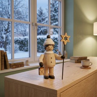 Miniatur „Mädchen mit Stern“ (ca. 5 cm) aus naturbelassenem Holz, mit kleiner Figur in Winterkleidung, die eine Laterne und einen Sternstab hält. Die Figur steht auf einem Holzmöbel in einer gemütlichen Stube; im Hintergrund ein Fenster mit verschneiter Winterlandschaft sowie Bücher und eine Tasse, die eine ruhige, winterliche Atmosphäre schaffen.