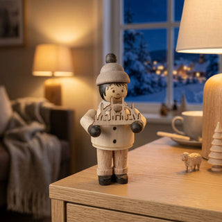 Miniatur „Junge mit Schwibbogen“ (ca. 5 cm) aus naturbelassenem Holz, mit kleiner Figur in Winterkleidung, die einen filigranen Schwibbogen mit winterlichem Motiv in den Händen hält. Die Figur steht auf einem Holzmöbel in einer gemütlichen Stube; im Hintergrund ein Fenster mit verschneiter Winterlandschaft, eine Lampe, eine Tasse und kleine Dekofiguren sorgen für eine warme, weihnachtliche Atmosphäre.