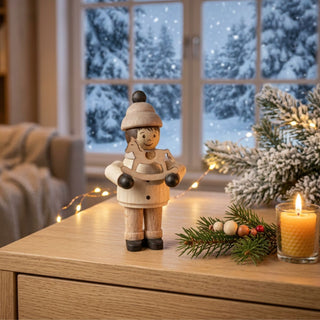 Miniatur „Junge mit Schaukelpferd“ (ca. 5 cm) aus naturbelassenem Holz, mit einer kleinen Figur in Winterkleidung, die ein filigranes Schaukelpferd in den Händen hält. Die Figur steht auf einem Holzmöbel in einer gemütlichen Wohnstube; im Hintergrund ein Fenster mit verschneiter Winterlandschaft, eine Kerze, Tannenzweige und Lichterkette schaffen eine warme, weihnachtliche Atmosphäre.
