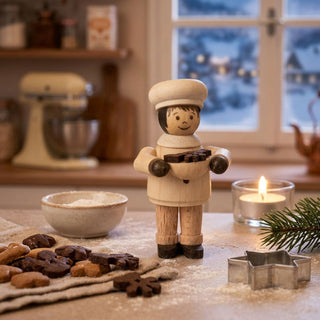 Miniatur „Bäckermädchen“ (ca. 5 cm) aus naturbelassenem Holz, mit kleiner Figur in Bäckerkleidung und Mütze, die eine Schale mit Gebäck hält. Die Figur steht auf einer bemehlten Arbeitsfläche in einer gemütlichen Küche; im Vordergrund Kekse und Ausstechformen, im Hintergrund ein Fenster mit verschneiter Winterlandschaft sowie eine brennende Kerze, die eine warme, weihnachtliche Atmosphäre schaffen.
