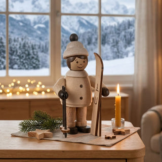 Miniatur „Mädchen mit Ski“ (ca. 5 cm) aus naturbelassenem Holz, mit kleiner Figur in Winterkleidung und Mütze, die ein Paar Ski und einen Skistock hält. Die Figur steht auf einem Holztisch in einer gemütlichen Wohnstube; im Hintergrund ein großes Fenster mit verschneiter Berglandschaft, eine Kerze und Lichter sorgen für eine warme, winterliche Atmosphäre.