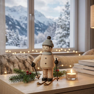Miniatur „Junge auf Ski“ (ca. 5 cm) aus naturbelassenem Holz, mit kleiner Figur in Winterkleidung und Mütze, die auf Skiern steht und Skistöcke hält. Die Figur steht auf einem Holztisch in einer gemütlichen Wohnstube; im Hintergrund ein großes Fenster mit verschneiter Berglandschaft, dazu eine Kerze, Bücher und eine Lichterkette, die eine warme, winterliche Atmosphäre schaffen.