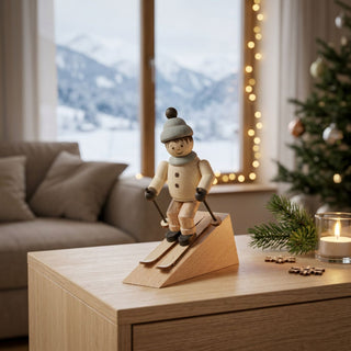 Miniatur „Abfahrtsläufer“ (ca. 5 cm) aus naturbelassenem Holz, mit kleiner Figur in Winterkleidung und Mütze, die auf Skiern eine Schräge hinabfährt und Skistöcke hält. Die Figur steht auf einem Holztisch in einer gemütlichen Wohnstube; im Hintergrund ein großes Fenster mit verschneiter Berglandschaft, dazu Lichterkette, Weihnachtsbaum, Tannenzweig und Kerze, die eine warme, winterliche Atmosphäre schaffen.