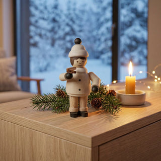 Miniatur „Mädchen mit Bratwurst“ (ca. 5 cm) aus naturbelassenem Holz, mit kleiner Figur in Winterkleidung und Mütze, die eine Bratwurst in der Hand hält. Die Figur steht auf einem Holztisch in einer gemütlichen Wohnstube; im Hintergrund ein Fenster mit verschneiter Winterlandschaft, Tannenzweig mit Zapfen, Kerze und Lichter sorgen für eine warme, winterliche Atmosphäre.