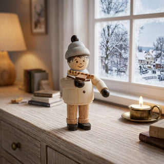 Miniatur „Junge mit Bratwurst“ (ca. 5 cm) aus naturbelassenem Holz, mit kleiner Figur in Winterkleidung und Mütze, die eine Bratwurst in der Hand hält. Die Figur steht auf einem Holzmöbel in einer gemütlichen Wohnstube; im Hintergrund ein Fenster mit verschneiter Winterlandschaft, Bücher und eine Kerze sorgen für eine warme, winterliche Atmosphäre.