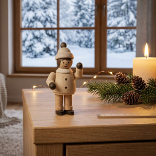 Miniatur „Mädchen winkend“ (ca. 5 cm) aus naturbelassenem Holz, mit kleiner Figur in Winterkleidung und Mütze, die eine Hand zum Winken hebt. Die Figur steht auf einem Holztisch in einer gemütlichen Wohnstube; im Hintergrund ein Fenster mit verschneiter Winterlandschaft, dazu eine Kerze, Tannenzweig mit Zapfen und eine Lichterkette, die eine warme, winterliche Atmosphäre schaffen.