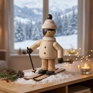 Miniatur „Mädchen auf Ski“ (ca. 5 cm) aus naturbelassenem Holz, mit kleiner Figur in Winterkleidung und Mütze, stehend auf Skiern und Skistöcke in den Händen haltend. Die Figur steht auf einer mit Kunstschnee dekorierten Holzfläche; im Hintergrund ein Fenster mit verschneiter Berglandschaft, dazu eine Kerze, Lichterkette und winterliche Dekoration mit Tannenzweig und Zapfen, die eine warme, gemütliche Atmosphäre schaffen.