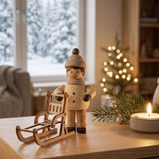 Miniatur „Mädchen mit Schlitten“ (ca. 5 cm) aus naturbelassenem Holz, mit kleiner Figur in Winterkleidung und Mütze, die neben einem filigranen Holzschlitten steht. Die Szene befindet sich auf einem Holztisch in einer gemütlichen Wohnstube; im Hintergrund ein Fenster mit verschneiter Winterlandschaft, dazu Weihnachtsbaum mit Lichtern, Kerze und Tannenzweig, die eine warme, festliche Atmosphäre schaffen.
