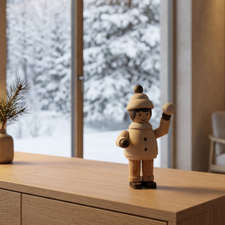 Miniatur „Mädchen mit Schneeball“ (ca. 5 cm) aus naturbelassenem Holz, mit kleiner Figur in Winterkleidung und Mütze, die einen Schneeball in der erhobenen Hand hält. Die Figur steht auf einem Holzmöbel vor einem großen Fenster mit verschneiter Winterlandschaft; im Hintergrund sind Bäume unter Schnee sowie dezente Wohnraumdekoration, die eine ruhige, winterliche Atmosphäre schaffen.