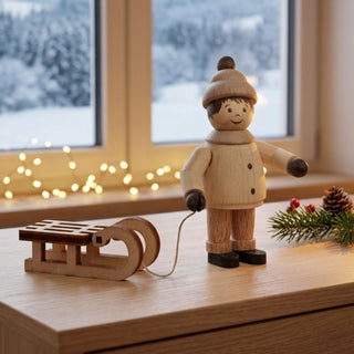 Miniatur „Junge mit Schlitten“ (ca. 5 cm) aus naturbelassenem Holz, mit kleiner Figur in Winterkleidung und Mütze, die einen Holzschlitten an einer Schnur hinter sich herzieht. Die Figur steht auf einem Holztisch in einer gemütlichen Wohnstube; im Hintergrund ein Fenster mit verschneiter Winterlandschaft sowie Lichterkette, Tannenzweig mit Zapfen und warmes Licht, die eine festliche Atmosphäre schaffen.