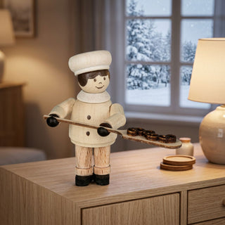 Miniatur „Brezelbäckerin“ (ca. 5 cm) aus naturbelassenem Holz, mit kleiner Figur in Bäckerkleidung und Mütze, die ein Backblech mit mehreren Brezeln hält. Die Figur steht auf einem Holzmöbel in einer gemütlichen Wohnstube; im Hintergrund ein Fenster mit verschneiter Winterlandschaft sowie eine Tischlampe und kleine Dekoelemente, die eine warme, winterliche Atmosphäre schaffen.