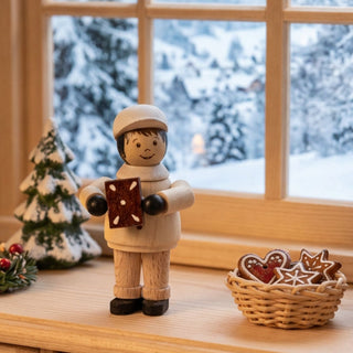Miniatur „Lebkuchenbäcker“ (ca. 5 cm) aus naturbelassenem Holz, mit kleiner Figur in Bäckerkleidung und Mütze, die ein Lebkuchenherz in den Händen hält. Die Figur steht auf einem Holzmöbel in einer gemütlichen Wohnstube; im Hintergrund ein Fenster mit verschneiter Winterlandschaft, daneben ein kleiner Tannenbaum sowie ein Korb mit Lebkuchen, die eine warme, weihnachtliche Atmosphäre schaffen.