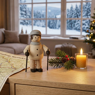 Miniatur „Nordic Walker“ (ca. 5 cm) aus naturbelassenem Holz, mit kleiner Figur in Winterkleidung und Mütze, die zwei Nordic-Walking-Stöcke hält. Die Figur steht auf einem Holztisch in einer gemütlichen Wohnstube; im Hintergrund ein großes Fenster mit verschneiter Berglandschaft, daneben eine brennende Kerze, eine Wanderkarte sowie Tannenzweig mit Zapfen, die eine ruhige, winterliche Atmosphäre schaffen.