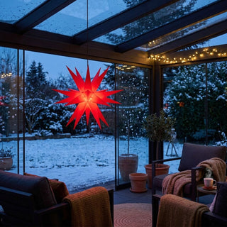 Roter LED-Adventsstern von SAICO mit 60 cm Durchmesser, leuchtend an einer Überdachung im Außenbereich aufgehängt. Der Stern strahlt warmes Licht aus und setzt einen festlichen Akzent in einem winterlichen Garten mit Schnee. Die gemütliche Terrasse ist mit Sitzmöbeln und einer Lichterkette dekoriert.