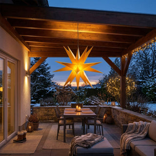 Gelber LED-Adventsstern für den Außenbereich von SAICO mit 60 cm Durchmesser, aufgehängt unter einer überdachten Terrasse. Der leuchtende Weihnachtsstern strahlt warmes Licht aus und schafft eine festliche Atmosphäre. Im Hintergrund sind ein winterlicher Garten mit Schnee, stimmungsvolle Lichterdekorationen sowie eine gemütlich eingerichtete Terrasse mit Esstisch und Sitzmöbeln zu sehen.