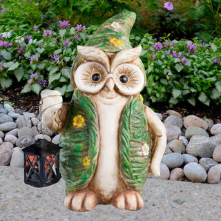 Dekorative Eulen-Gartenfigur mit grünem Hut und grünem Umhang, verziert mit kleinen Blumen. Die Eule hält eine kleine schwarze Laterne und steht auf einem Steinweg vor üppigem grünen Pflanzenbeet mit violetten Blüten.
