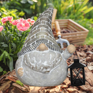 Dekorativer Gartenwichtel aus Steinoptik mit hoher spitzer Mütze im Ziegelmuster, grauem Bart und runder Nase. Der Wichtel hält eine kleine schwarze Laterne und steht auf Rindenmulch, umgeben von grünen Pflanzen und rosa Blüten; im Hintergrund liegen Gartenwerkzeug und Holzkiste.