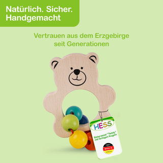 Ein hölzernes Babyspielzeug in Form eines Teddybären mit bunten Holzkugeln. Oben links im Bild befindet sich ein grünes Feld mit dem Text: „Natürlich. Sicher. Handgemacht“. Darunter steht: „Vertrauen aus dem Erzgebirge seit Generationen“. Am Spielzeug hängt ein Etikett mit der Aufschrift: „HESS Babyrassel 'Teddy' mit farbigen Kugeln 100% MADE IN GERMANY“.