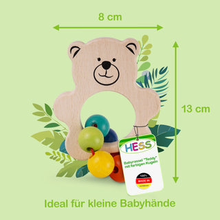 Eine hölzerne Baby-Rassel in Form eines lächelnden Teddybären mit bunten Holzkugeln (grün, blau, gelb und rot) an der Unterseite. Die Rassel ist etwa 8 cm breit und 13 cm hoch. Im Hintergrund sind grüne Blätter zu sehen. Ein Etikett an der Rassel zeigt den Schriftzug „HESS“ sowie „Babyrassel 'Teddy' mit farbigen Kugeln“ und „100% MADE IN GERMANY“. Am unteren Bildrand steht: „Ideal für kleine Babyhände“.