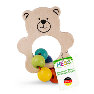 Ein hölzerner Babygreifling in Form eines lächelnden Teddybären mit einem runden Loch in der Mitte. Am unteren Teil sind vier bunte Holzkugeln (grün, gelb, blau und rot) befestigt. Der Bär hat ein gezeichnetes Gesicht mit schwarzen Augen, Nase und Mund. Ein Etikett am Greifling trägt die Aufschrift: „HESS Babyrassel 'Teddy' mit farbigen Kugeln 100% MADE IN GERMANY.“