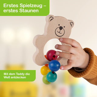 Ein Kind hält ein hölzernes Spielzeug in Form eines Teddybären mit lachendem Gesicht und einem Loch in der Mitte. Durch das Loch hängen bunte Holzkugeln an einer Schnur. Im Bild stehen zwei Texte: oben links „Erstes Spielzeug – erstes Staunen“ und unten links „Mit dem Teddy die Welt entdecken“.