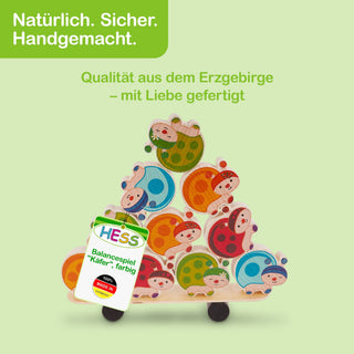 „Ein Holzspielzeug in Form eines Balancespiels mit bunten Marienkäfern, die zu einer Pyramide gestapelt sind. Der Hintergrund ist hellgrün. Oben links steht der Text ‚Natürlich. Sicher. Handgemacht.‘ Unter dem Spielzeug steht ‚Qualität aus dem Erzgebirge – mit Liebe gefertigt.‘ Ein Etikett am Spielzeug trägt die Aufschrift ‚HESS Balancespiel „Käfer“, farbig‘ sowie ‚100 % MADE IN GERMANY‘.“
