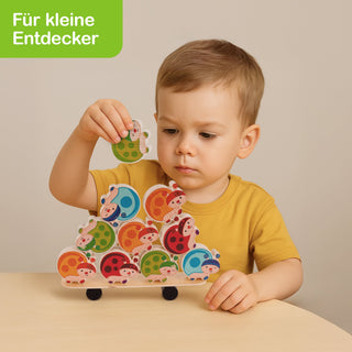 „Ein Kind mit gelbem Shirt spielt mit einem Stapelspiel aus bunten, marienkäferförmigen Holzstücken. Das Gesicht des Kindes ist unscharf dargestellt. Es hält ein Teil in der Hand und setzt es oben auf den Stapel. In der oberen linken Ecke steht der grüne Text ‚Für kleine Entdecker‘.“