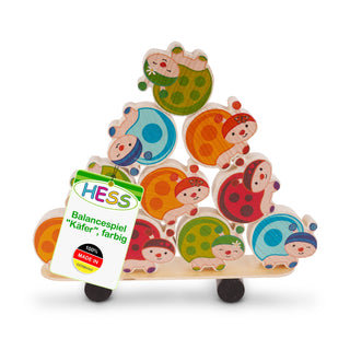 „Holzspielzeug in Form eines Balancespiels mit bunten Marienkäfern, die zu einer Pyramide gestapelt sind. Die Marienkäfer sind in den Farben Grün, Blau, Orange, Rot und Gelb bemalt, haben lächelnde Gesichter und Punkte auf dem Rücken. Ein Etikett am Spielzeug trägt die Aufschrift ‚HESS Balancespiel „Käfer“, farbig‘ sowie ‚100 % MADE IN Germany‘.“