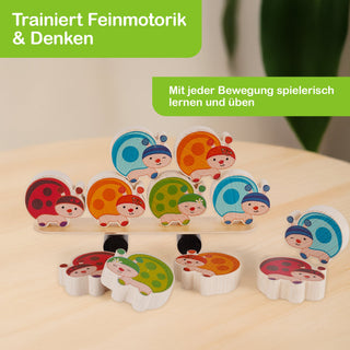 „Sechs bunte Holz-Marienkäfer liegen und stehen auf einer hellen Oberfläche. Die Käfer sind in Rot, Orange, Grün und Blau bemalt. Einige sind gestapelt, andere einzeln platziert. Der Hintergrund ist unscharf und zeigt eine Innenraumszene mit etwas Grün. Im Bild steht der Text: ‚Trainiert Feinmotorik & Denken‘ sowie ‚Mit jeder Bewegung spielerisch lernen und üben‘.“