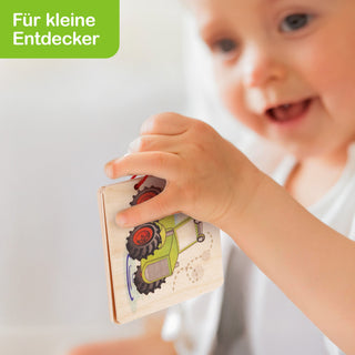 „Ein Kind hält ein kleines Holz-Bilderbuch, auf dessen Seite ein grüner Traktor abgebildet ist. In der oberen linken Ecke befindet sich ein grünes Feld mit weißer Schrift: ‚Für kleine Entdecker‘.“
