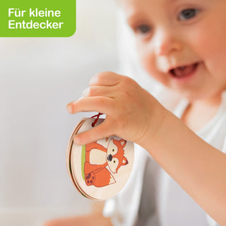 „Ein Kind hält ein rundes Holzbuch mit der Illustration eines Fuchses. Oben links im Bild steht in weißer Schrift auf grünem Hintergrund: ‚Für kleine Entdecker‘.“